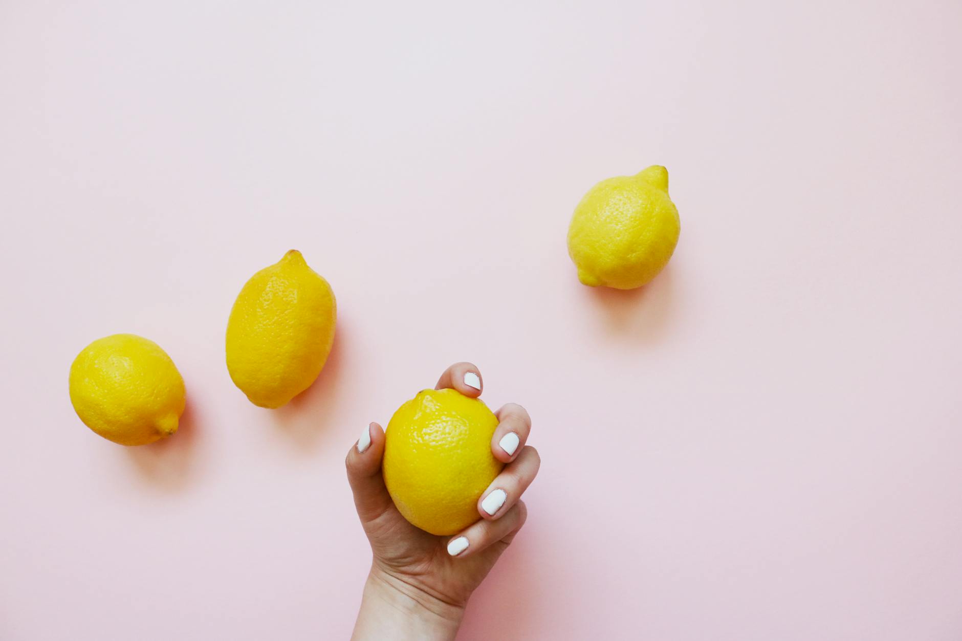 Una mano sosteniendo un limón fresco sobre fondo rosa pastel rodeado de limones adicionales