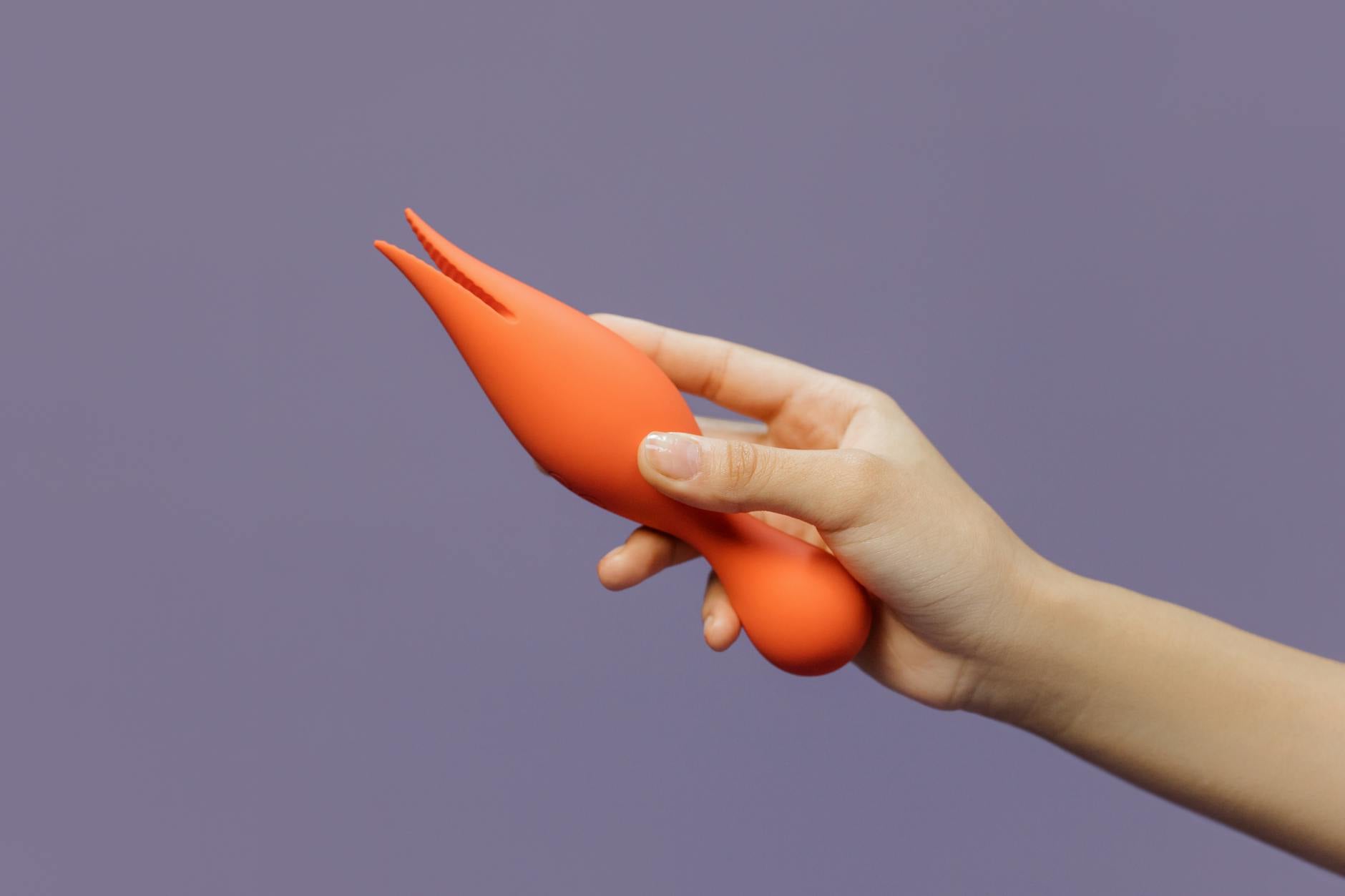 Primer plano de una mano sosteniendo un vibrador naranja contra un fondo púrpura minimalista, mostrando sensualidad moderna.