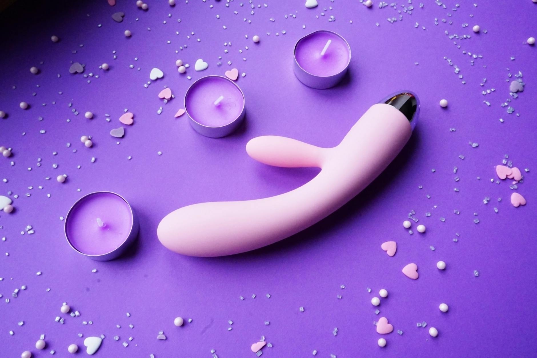 Vibrador clitoral rosa sobre fondo púrpura con confeti en forma de corazón y velas encendidas para crear un ambiente romántico